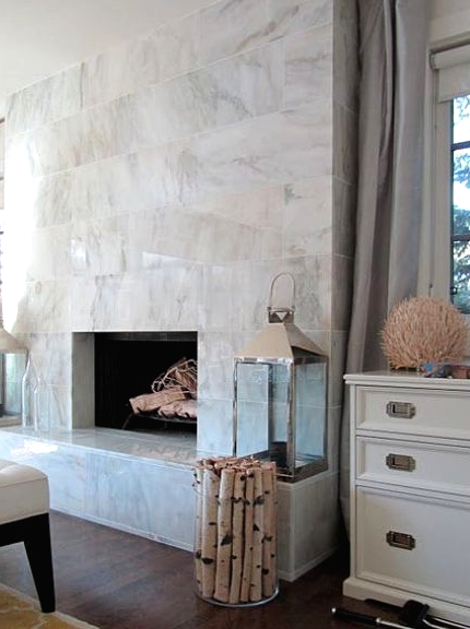 a large non-working fireplace clad with white marble tiles all over, with candle lanterns and firewood in a vase