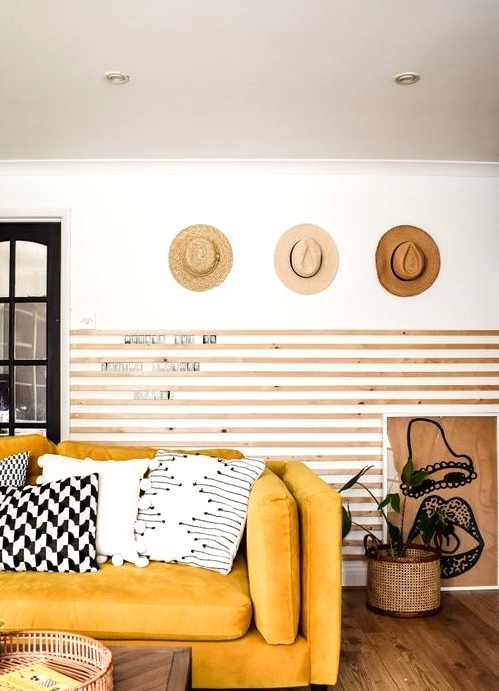 a chic living room with wooden slabs on the wall, a yellow sofa and graphic pillows, hats on display and some plants
