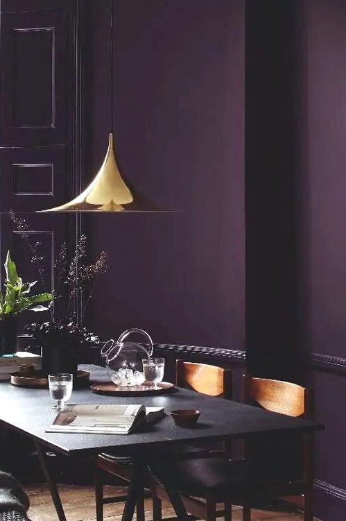 a moody dining room with an aubergine accent wall, a black trestle table and wooden chairs, a shiny gilded pendant lamp