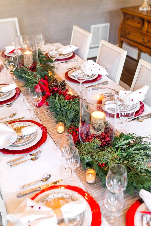 a bright and chic Christmas table setting with evergreens, red berries, candles wrapped with bark, red chargers and polka dot napkins