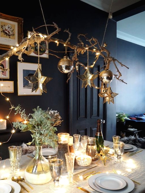 a chic Christmas tablescape with a branch with ornaments and stars hanging over the table, lights and greenery on the table and gold cutlery