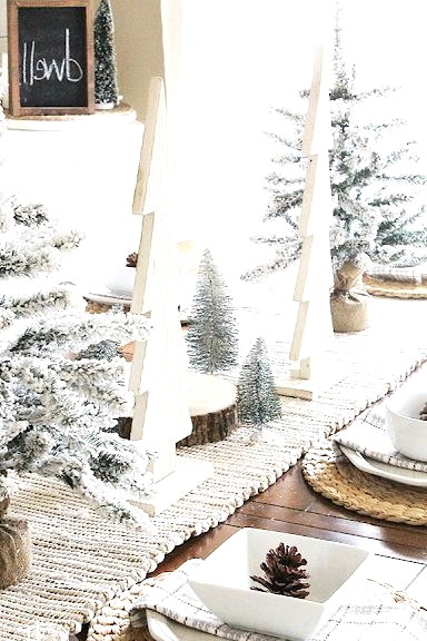 a farmhouse Christmas tablescape with a woven table runner and placemats, flocked and plywood Christmas trees, white porcelain and pinecones