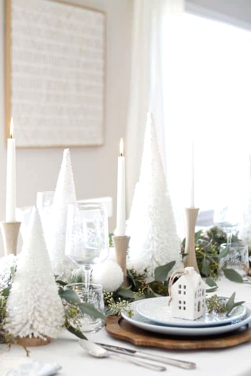 a lovely Christmas tablescape with white bottle brush trees, white candles in wooden candleholders, wooden placemats, white houses and fresh eucalyptus