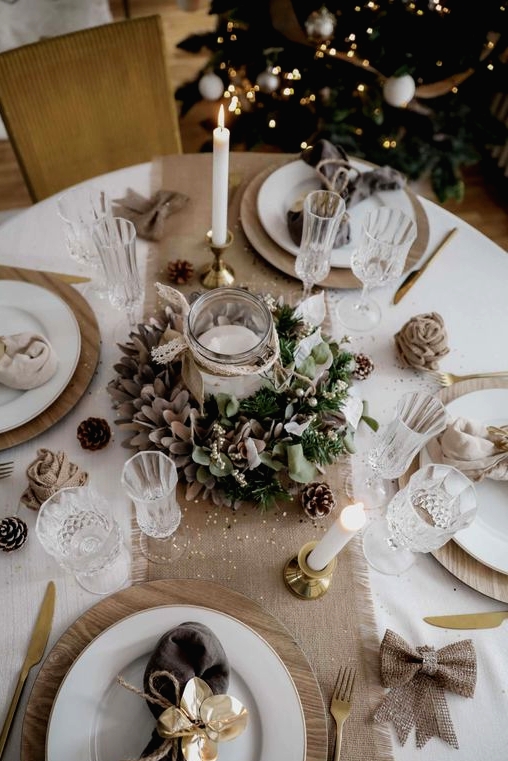 a neutral holiday tablescape with a burlap runner and bows, brown and neutral napkins, fabric blooms and greenery and pinecones