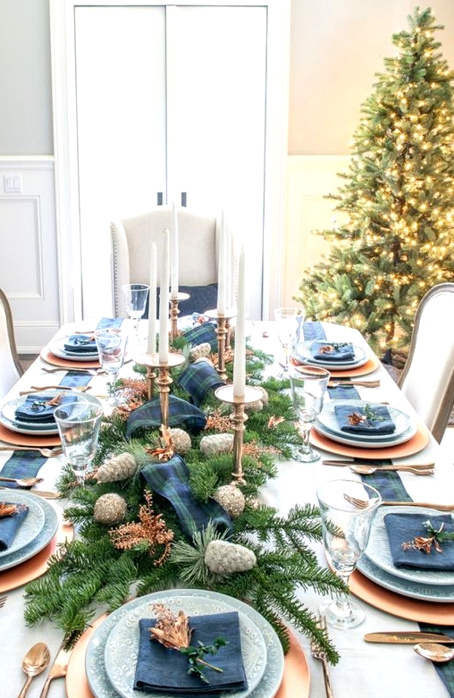 a pretty Christmas tablescape done in navy and copper, with tall and thin candles, printed plates, an evergreen and pinecone runner plus a plaid ribbon