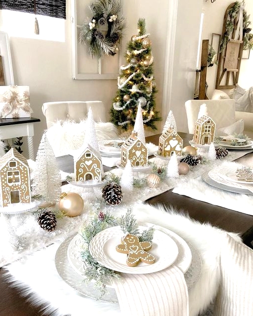 a super cute Christmas tablescape with a white faux snow table runner and faux fur placemats, snowy pinecones, gold ornaments, white bottle brush trees and gingerbread houses