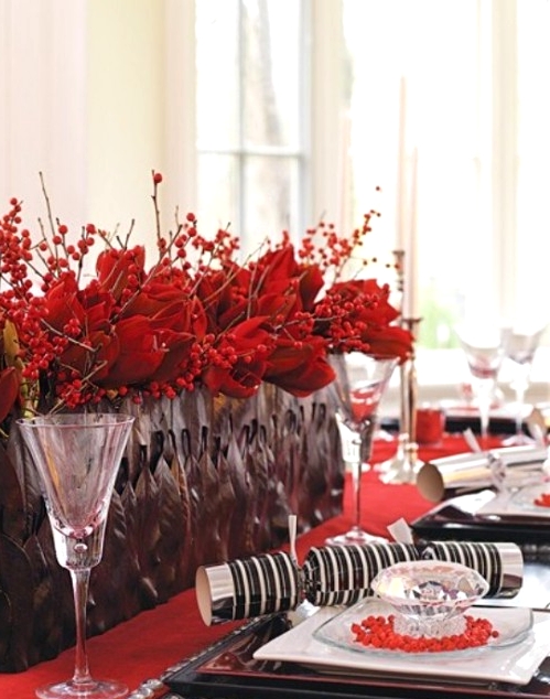 a bright Christmas tablescape with a red table runner, a black planter with red berries and poinsettias, elegant black square plates and chic glasses