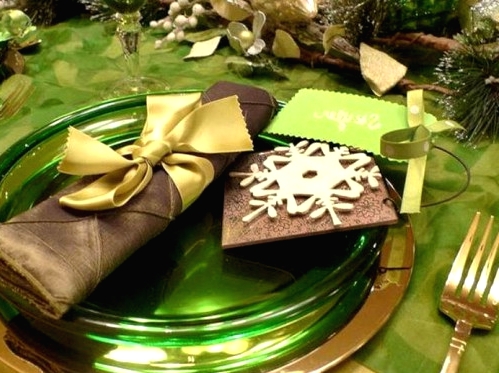 a chic grene, brown and gold Christmas tablescape with glass plates, a brown napkin and an envelope with a snowflake, green ribbons