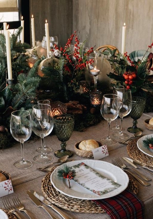 a fabulous rustic Christmas tablescape with lush evergreens, berries, candles, green glasses, woden placemats, red plaid napkins