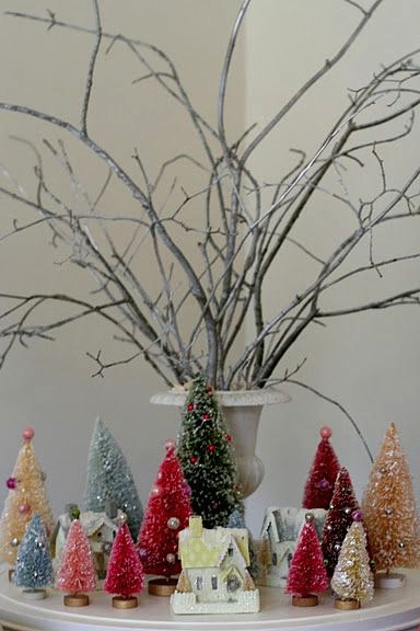 a small and colorful holiday arrangement with bright bottle brush trees and mini houses plus branches ina vintage urn