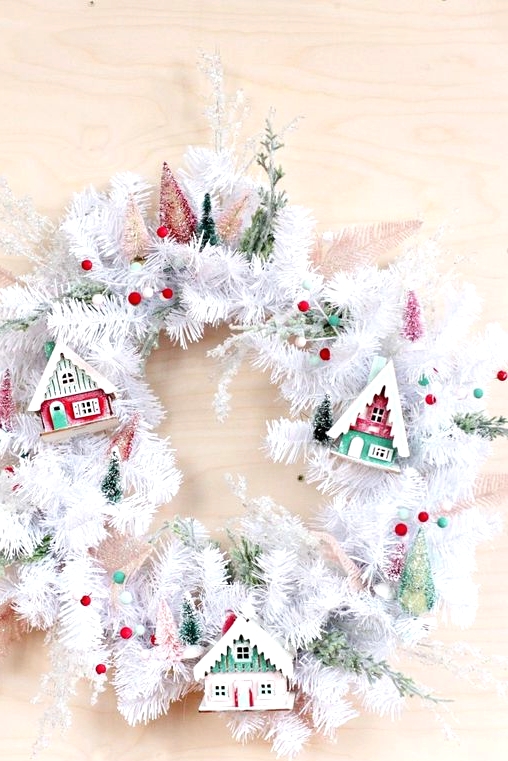 a white evergreen Christmas wreath with colorful pompoms, colorful small houses and bottle brush Christmas trees