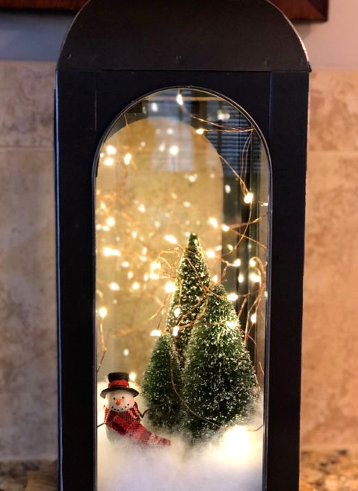 a black metal lantern with faux snow, bottle brush Christmas trees, lights and a small snowman is a lovely idea