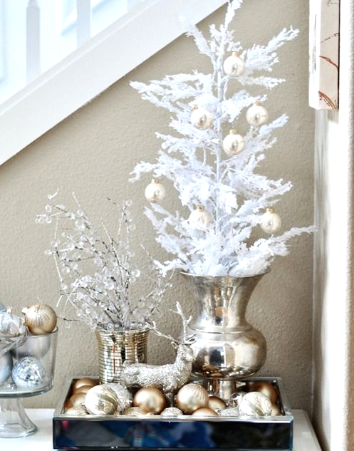 a gold urn with a white Christmas tree with gold ornaments, a tray with gold and gold glitter ornaments, a bowl with them and icy branches