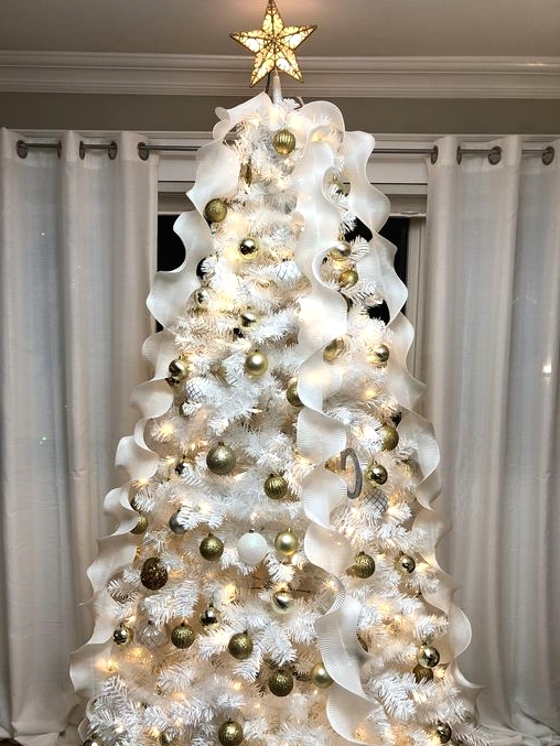 a white Christmas tree with gold and white ornaments, white ruffle ribbons and a lit up tree topper