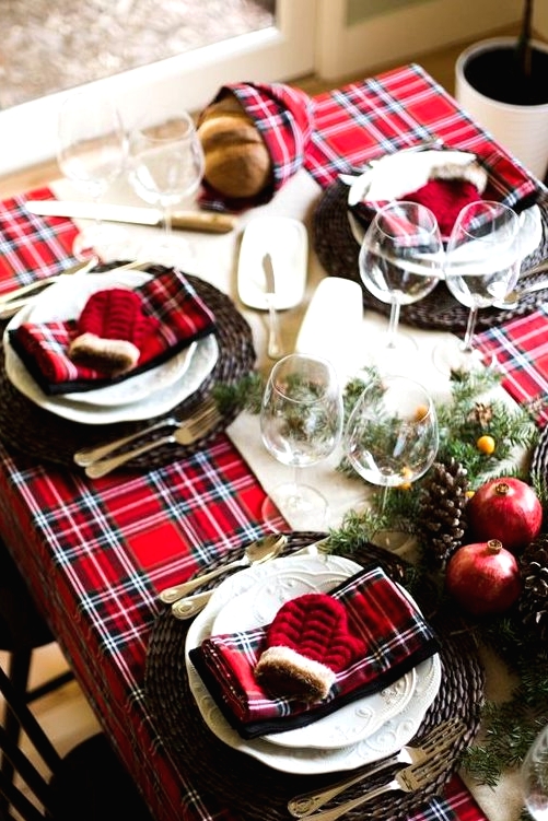 a bold Christmas table setting with a red plaid tablecloth and napkins, woven chargers, white porcelain, fir branches, berries, pomegranates and pinecones