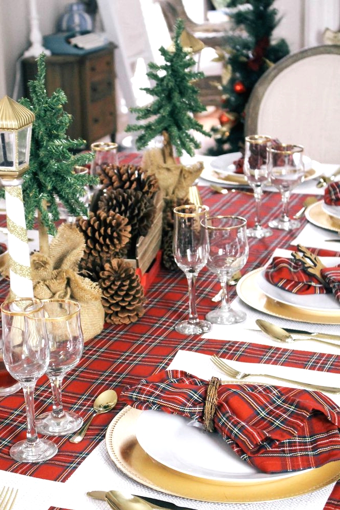 a lovely rustic Christmas tablescape with red plaid linens, gold chargers and cutlery, pinecones and mini fur trees wrapped in burlap
