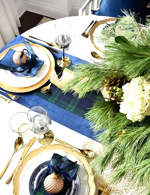 a refined Christmas tablescape with dark plaid linens, gold chargers and cutlery and neutral blooms and evergreens