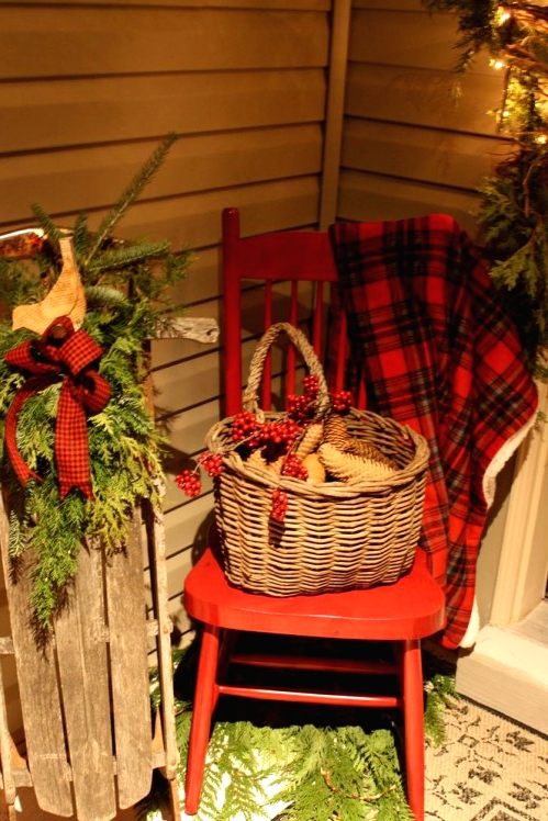 cozy Christmas porch decor with a red chair with a plaid blanket, a basket with pinecones and berries, a sleigh with evergreens and a red plaid bow, lights and branches is awesome