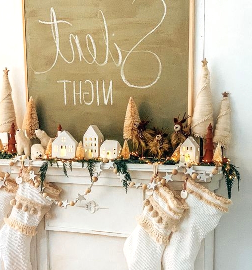 a holiday mantel with white tasseled stockings, an evergreen and light garland, stars and wooden beads, small houses and Christmas trees