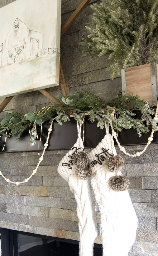 a pretty modern farmhouse mantel with an evergreen garland, wooden beads, white stockings and pompoms is amazing