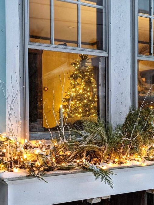 an outdoor window box with evergreens, vine, pinecones and lights is a cool way to the exterior of your house for Christmas
