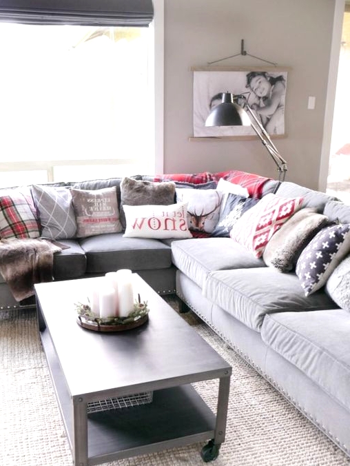a Christmassy living room with a grey sectional, lots of grey and red printed pillows, a grey table with pillar candles and a lamp