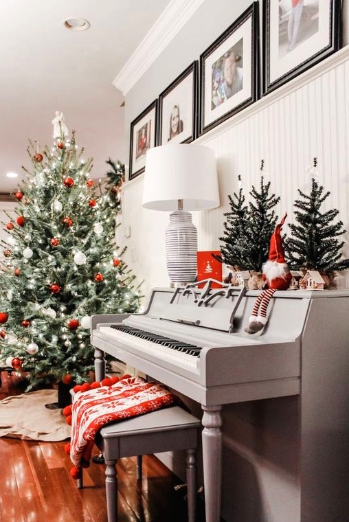 a vintage grey piano with some Christmas trees, a gnome, a grey bench with a printed red blanket, a Christmas tree with red and white ornaments