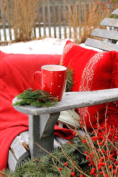 a simple wooden grey chair styled with a bold red blanket and a pillow, some evergreens with berries and a red mug is a great piece to enjoy winter outdoors