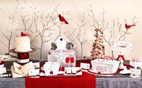 a catchy Christmas sweets table with a grey tablecloth, red runners and beads, white sweets and mugs and some red birds sitting on the branches over the table