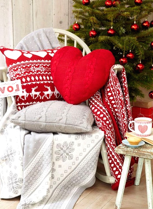 a chair styled for Christmas with knit pillows, grey and red printed blankets and pillows, a Christmas tree with red ornaments in the backdrop
