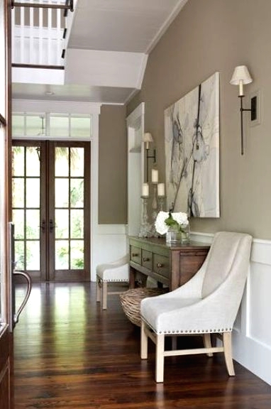 a vintage greige entryway with white wainscoting, a stained console table, white chairs, a basket, an oversized artwork and sconces