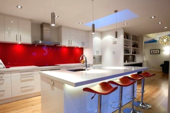 a modern white kitchen with sleek cabinets, a hot red glass backsplash, a lit up kitchen island and orange stools for a touch of color