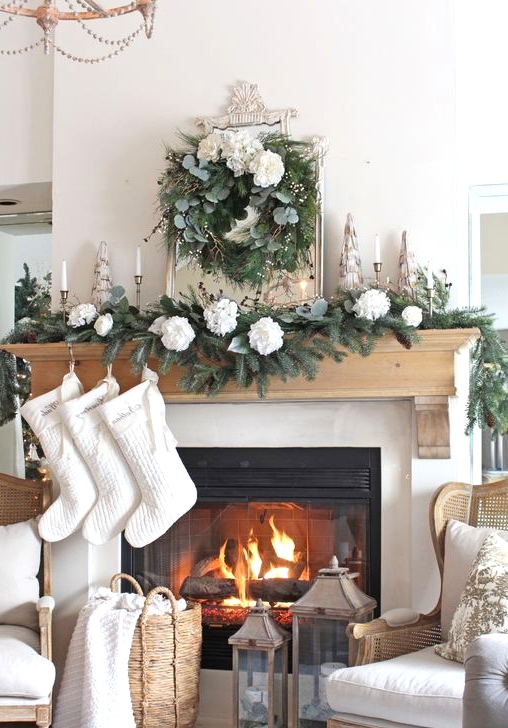 a chic Christmas mantel with an evergreen and white flower garland, candles, a mirror with a greenery and whie bloom wreath plus white stockings