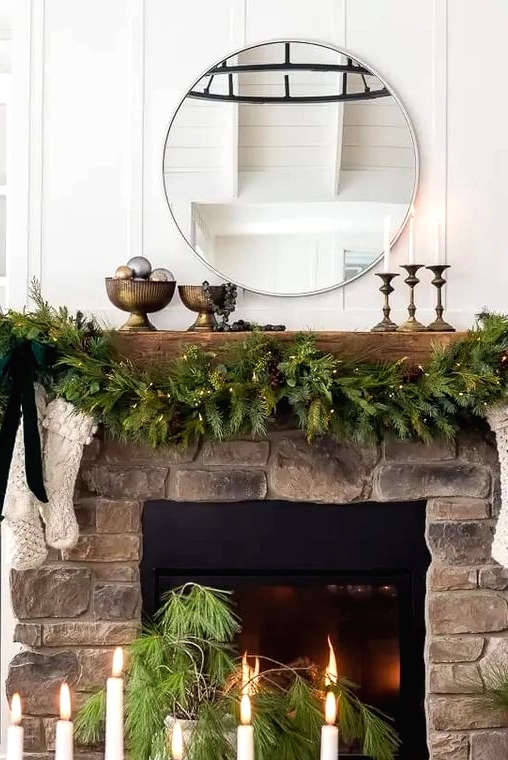 a Christmas mantel with an evergreen and light garland with pinecones, white stockings and a green bow, candles in candleholders