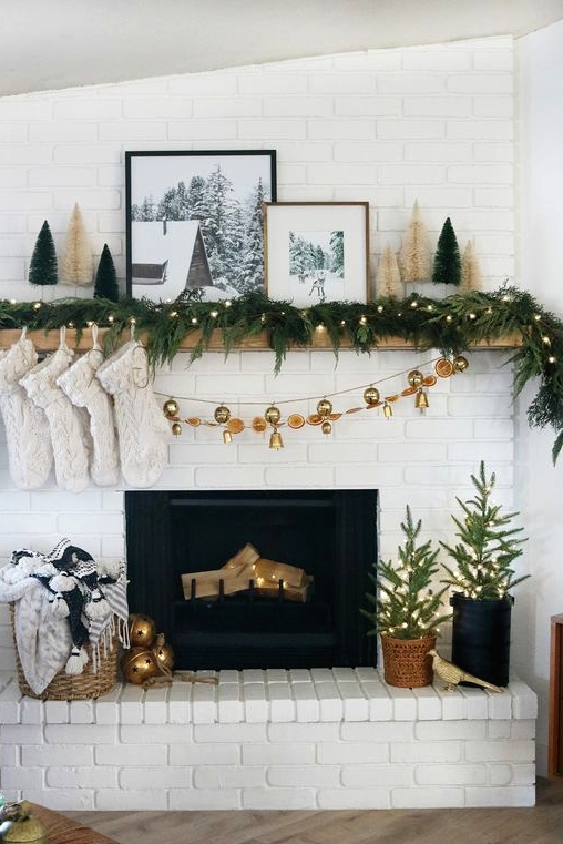 a modern glam Christmas mantel with an evergreen and light garland, white stockings, mini bottle cleaner trees and winter photos plus a bell garland