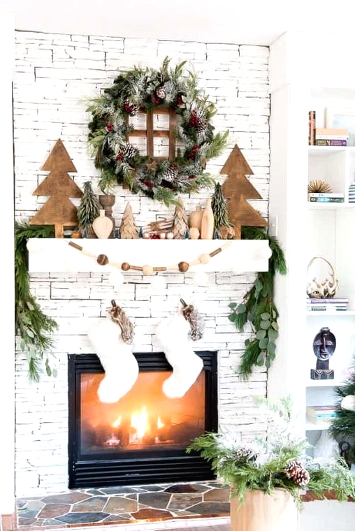 a woodland Christmas mantel with wooden Christmas trees, vases, wooden beads, an evergreen wreath and a garland plus white stockings