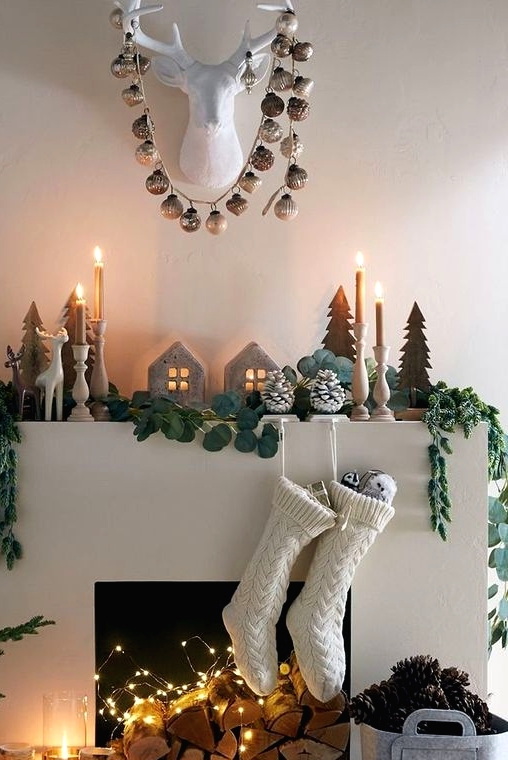 an elegant Scandinavian Christmas mantel with wooden Christmas trees, a greenery garland, candles in wooden candleholders, mini houses and snowy pinecones