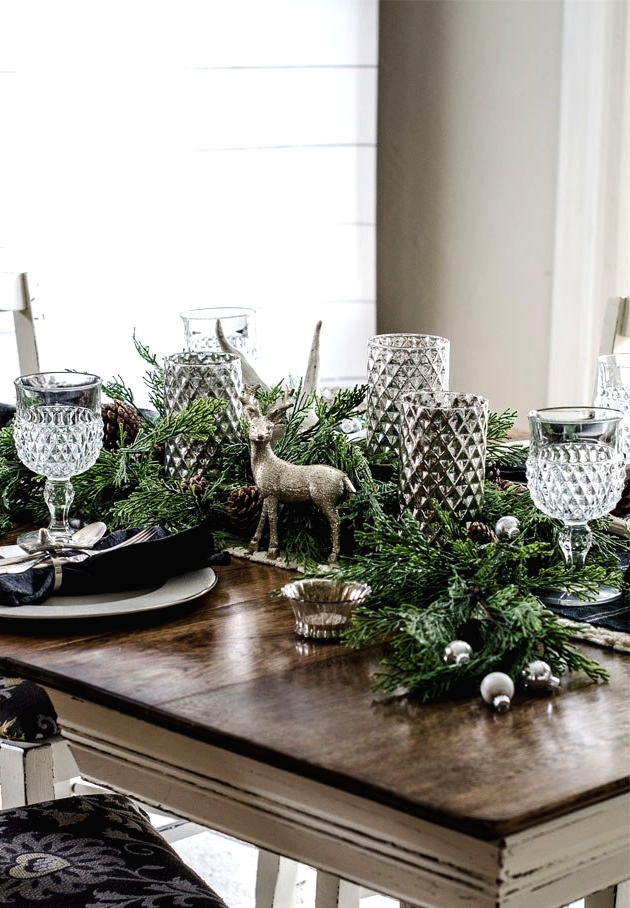 a forest glam Christmas tablescape with a greenery runner, silver ornaments, pinecones, glam and shiny candleholders, elegant glasses, dark napkins and silver chargers