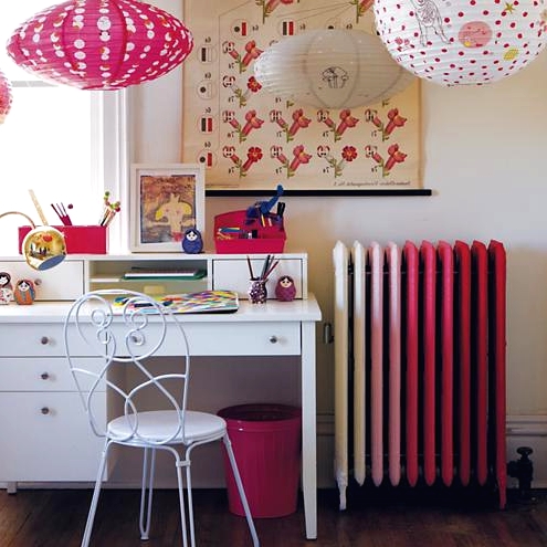 a bright space with an ombre pink radiator, a white desk and a chair, paper pendant lamps and bold artworks plus a bright rug