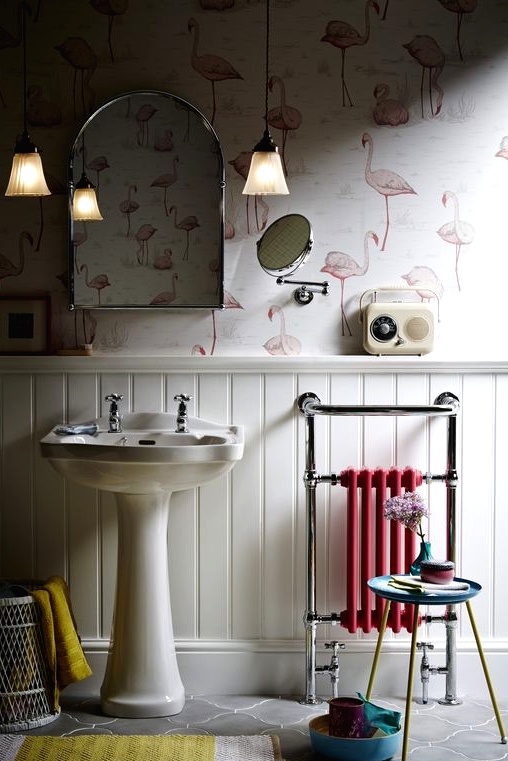 a lovely bathroom with flamingo printed wallpaper and white wainscoting, a free-standing sink, a small pink radiator, a bold rug and pendant lamps