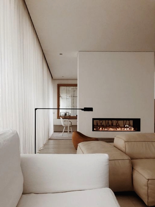 a neutral minimalist living room with double-sided fireplace, a tan leather sofa, a white one and an orcher chair by the fireplace