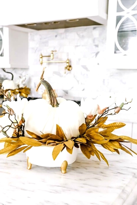 a glam Thanksgiving decoration of a white pumpkins, white blooms, gilded leaves on a white and gold stand is wow