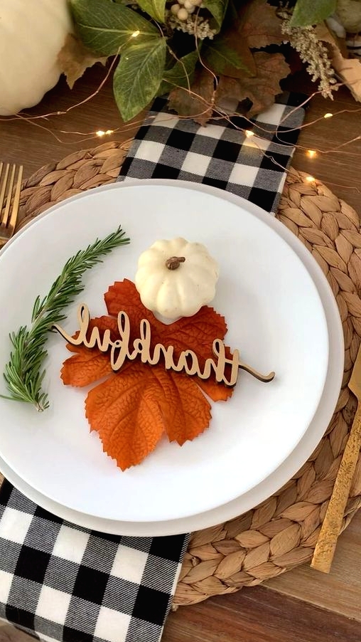 a pretty modern farmhouse tablescape with a woven placemat, white porcelain, a fall leaf, a pumpkin, greenery and calligraphy plus lights