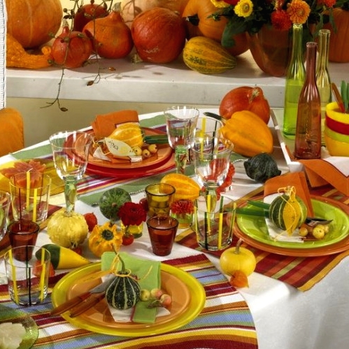 a colorful Thanksgiving tablescape with bold striped placemats, colored glasses, bottles and candleholders, some gourds and pumpkins is amazing