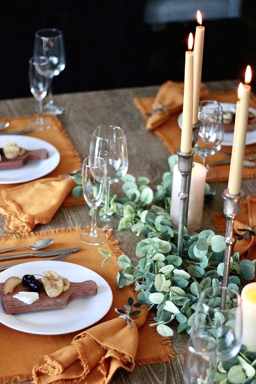 a delicate Thanksgiving place setting with a rust-colored placemat and a matching napkin, a white plate and a wooden board with some appetizers