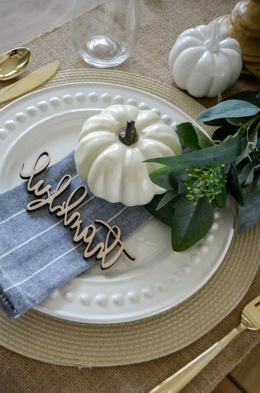 a pretty and natural Thanksgiving place setting with a woven charger, a plate, a faux pumpkin, a blue napkin and greenery