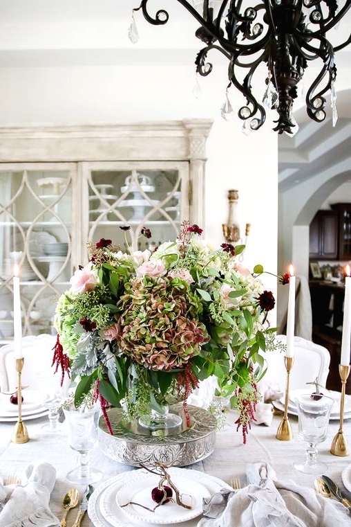 a gorgeous and lush Thanksgiving centerpiece of a bowl with green, blush and burgundy blooms on s tand is a sophisticated decor statement