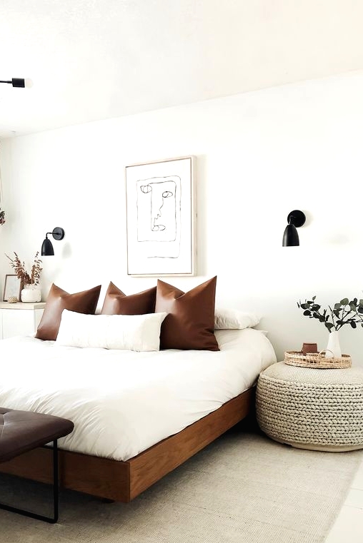 a chic mid-century modern bedroom in neutrals, with a rich-stained bed with neutral bedding, a leather bench, a woven ottoman and black sconces