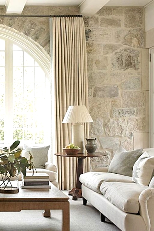 a French farmhouse living room in neutrals with a large arched French window, neutral furniture, a low coffee table and greenery