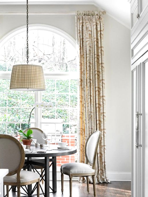a lovely dining room with an oversized French window, a round table and rounded chairs, a pendant lamp and printed curtains